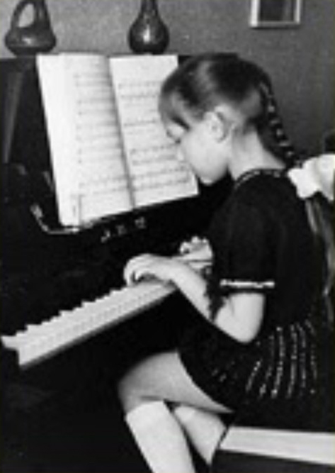 Katya Grineva as a young girl in Soviet Russia, seated in profile at an upright piano, reading sheet music with her hands on the keys, circa 1977