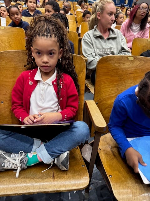 A young student in a red cardigan sits in a school auditorium, notebook in her lap, at a Katya Concerts for Kids performance
