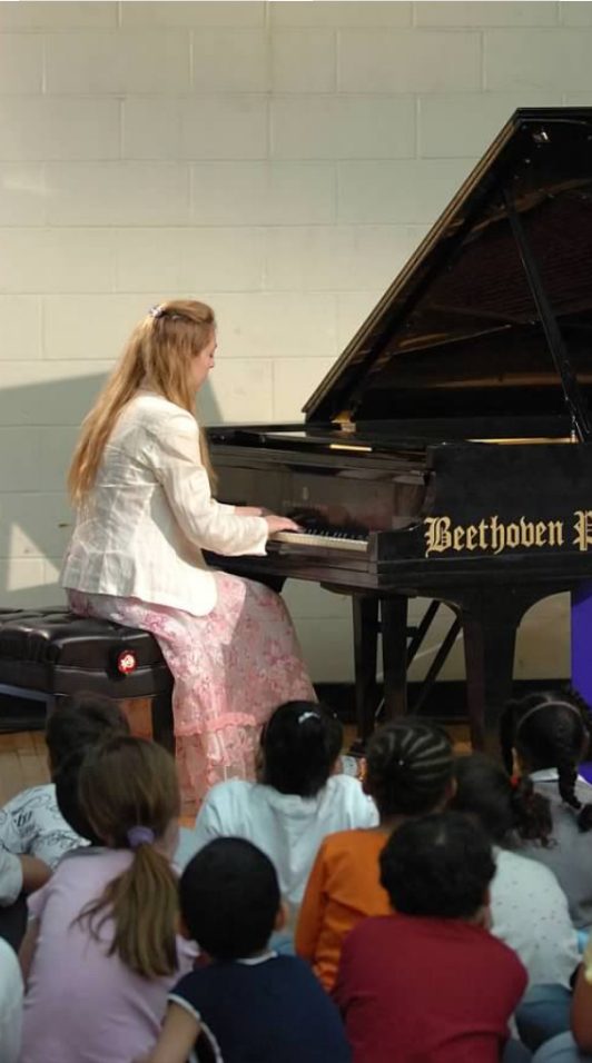 Katya Grineva plays a grand piano in a school gymnasium as a circle of children sit on the floor watching